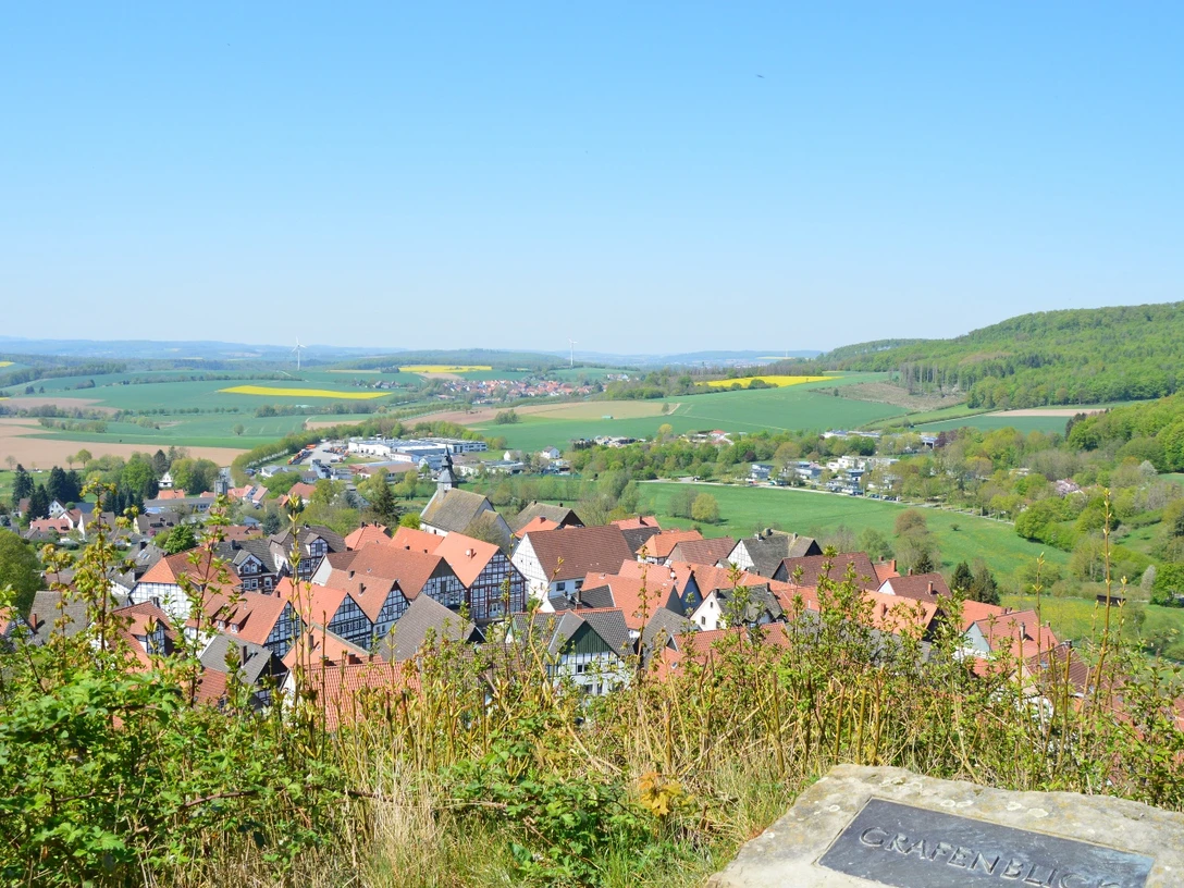 Grafenblick Blick auf ein Dorf mit roten Dächern, umgeben von grünen Hügeln und Feldern unter klarem Himmel.