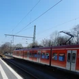 Ein roter Regionalzug steht am Gleis eines kleinen Bahnhofs, umgeben von Bäumen und Laternen.
