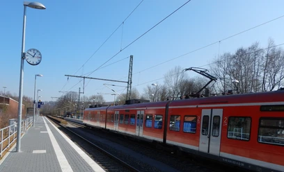 Bahnhof Ein roter Regionalzug steht am Gleis eines kleinen Bahnhofs, umgeben von Bäumen und Laternen.
