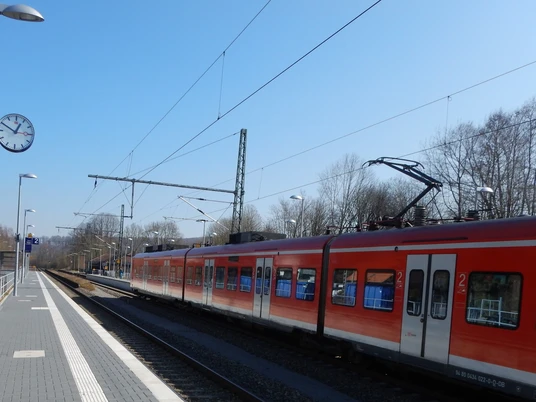 Bahnhof Ein roter Regionalzug steht am Gleis eines kleinen Bahnhofs, umgeben von Bäumen und Laternen.