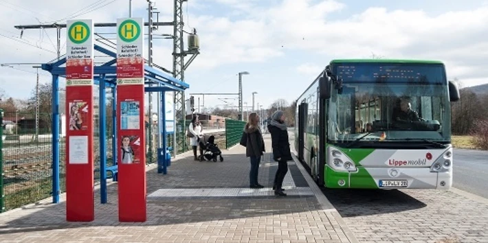 Bahnhof mit Busbahnhof Ein grüner Linienbus hält an einem Busbahnhof. Menschen steigen ein oder warten unter einem überdachten Bereich.
