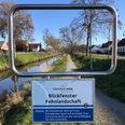 Treidelpfad mit Blickfenster Ein rechteckiges Stahlrahmenfenster zeigt einen Kanalweg. Dahinter liegen Häuser und Bäume. Ein Schild nennt den Blick auf eine Fehnlandschaft.
