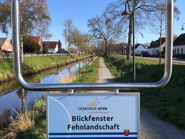 Treidelpfad mit Blickfenster Ein rechteckiges Stahlrahmenfenster zeigt einen Kanalweg. Dahinter liegen Häuser und Bäume. Ein Schild nennt den Blick auf eine Fehnlandschaft.
