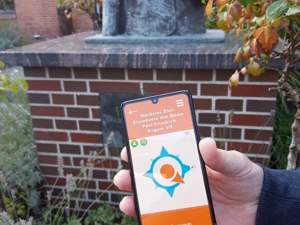 Wunderline Go App Eisenhüttenplatz Ein Handy zeigt die Karte der Wunderline Go App. Im Hintergrund ist ein Garten mit einer Skulptur zu sehen.