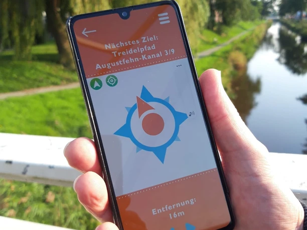 Wunderline Go App Treidelpfad Eine Hand hält ein Smartphone mit geöffneter Navigations-App vor dem Hintergrund eines sonnigen Kanals und grüner Landschaft.