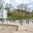 Flussbegegnungspunkt4.jpg Informationsschild und Sitzgelegenheiten an einem Flussufer mit angrenzendem Park und historischen Gebäuden.