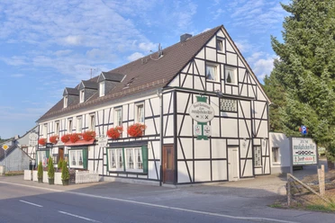 Lindlar - Hartegasse Gaststätte "Zum musikalischen Wirt" Tix Fachwerkhaus mit roten Blumenkästen, weißen Wänden, Schildern, blauem Himmel und Parkplatz daneben.