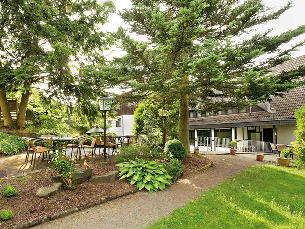 Hotel Goldener Acker Außenbereich eines Gasthauses mit Tischen unter Bäumen, umgeben von üppigem Grün und Pflanzen.