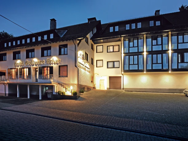 Hotel Goldener Acker Hotelgebäude mit beleuchteter Fassade und großer Fensterfront am Abend, parkender PKW vorne.