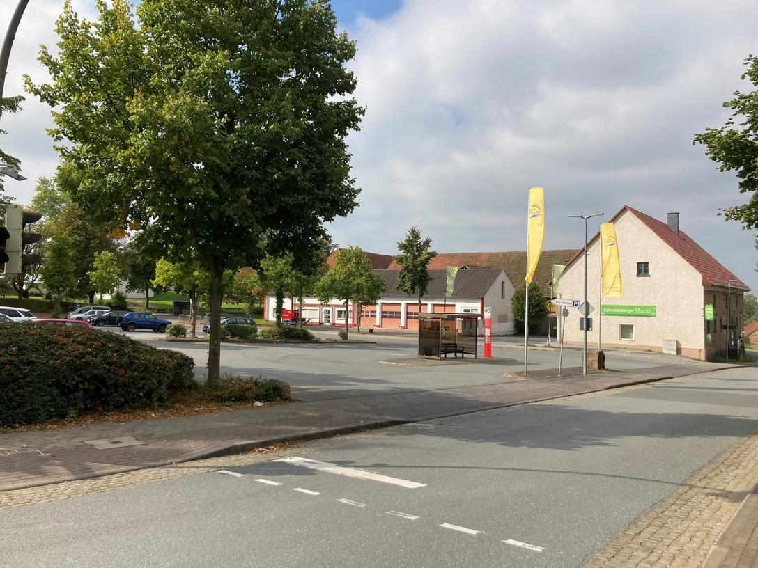 Parkplatz an einer ruhigen, grünen Straße mit Gebäuden. Einige Autos und Bäume im Hintergrund.