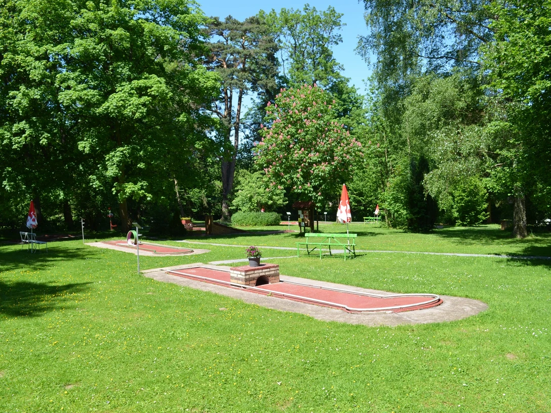 Minigolfplatz eingebettet in eine grüne Parklandschaft mit roten Bahnen und angrenzenden Bäumen.