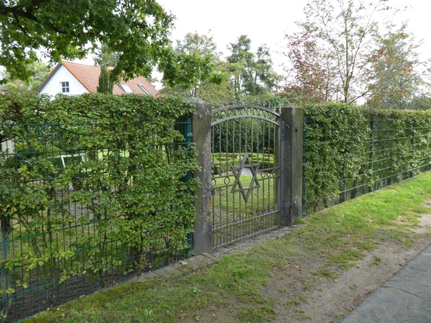 A metal gate bearing a Star of David leads to a Jewish cemetery surrounded by trees in a rural setting.