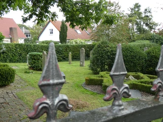 Jüdischer Friedhof Achim Eisenzaun mit Lilien verziert, im Hintergrund alte Grabsteine und umgebende Hecken auf grünem Gelände.