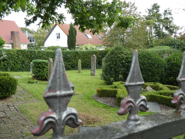 Jüdischer Friedhof Achim Eisenzaun mit Lilien verziert, im Hintergrund alte Grabsteine und umgebende Hecken auf grünem Gelände.