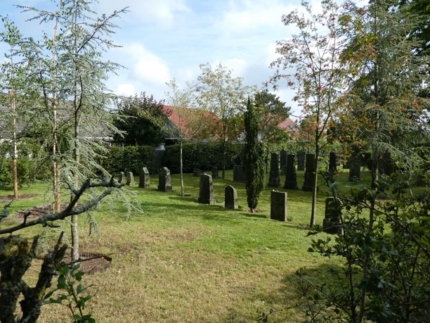 Kleine, historische Grabsteine auf dem grünen Jüdischen Friedhof Achim, umgeben von Bäumen und Büschen.
