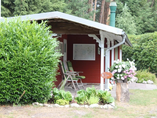 Ein rotes Holzgebäude mit überdachter Veranda, zwei hellgrünen Stühlen und blühenden Pflanzen.
