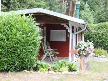 Moorcamp Ein rotes Holzgebäude mit überdachter Veranda, zwei hellgrünen Stühlen und blühenden Pflanzen.