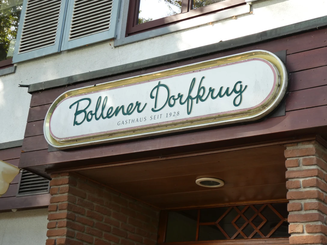Bollener Dorfkrug Weißes Schild mit grünem Schriftzug "Bollener Dorfkrug", Gasthaus seit 1928, an roter Backsteinfassade.