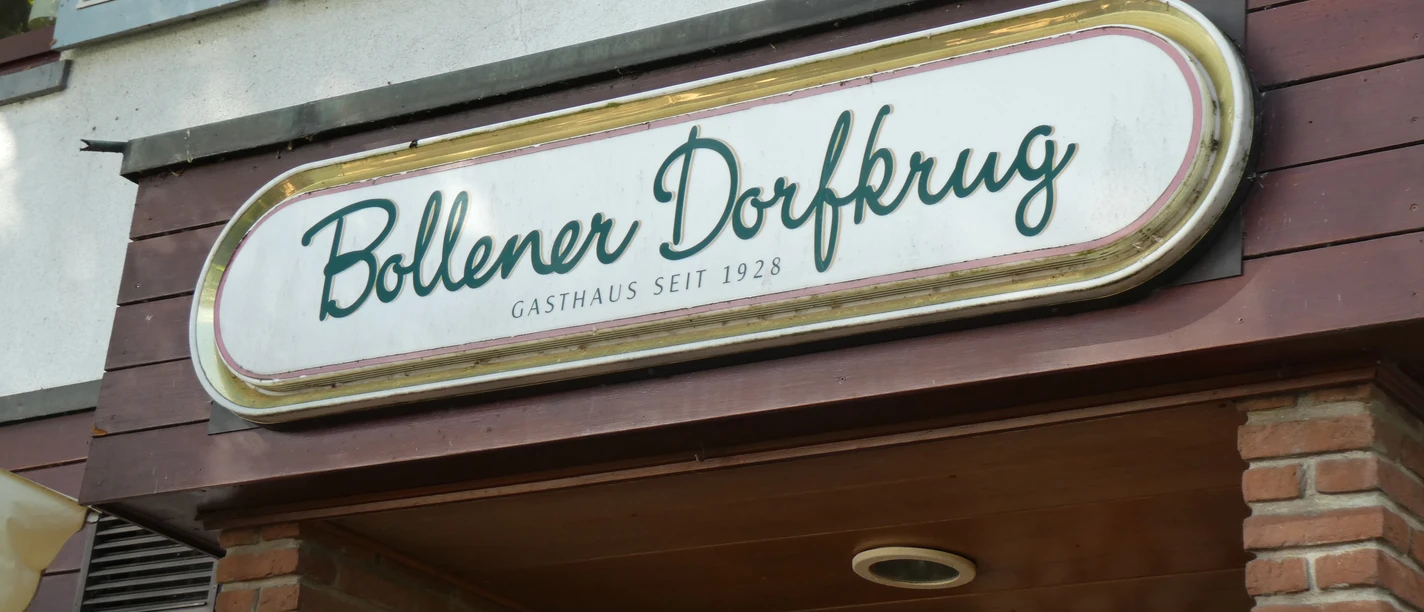Bollener Dorfkrug Weißes Schild mit grünem Schriftzug "Bollener Dorfkrug", Gasthaus seit 1928, an roter Backsteinfassade.