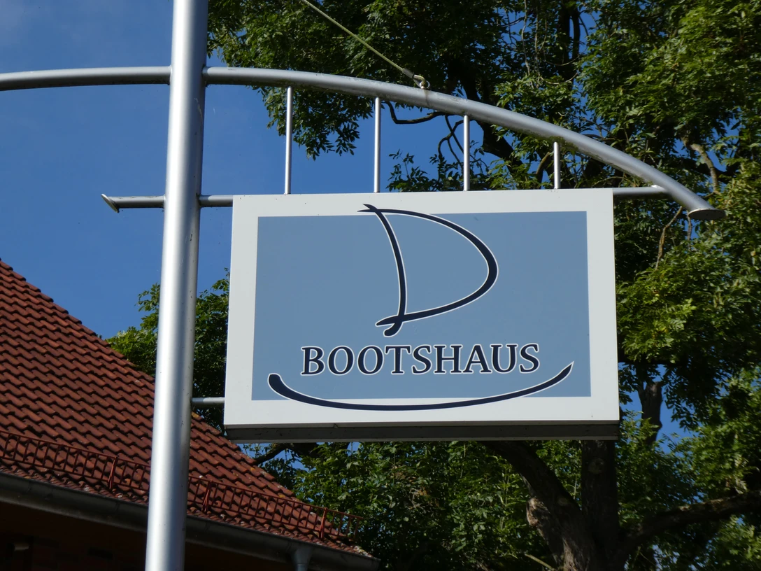 Holzschild mit segelbootförmigem Logo des Bootshaus Hotel-Restaurants vor grünen Bäumen.Wooden sign with sailboat-shaped logo of the Bootshaus Hotel-Restaurant in front of green trees.Træskilt med sejlbådsformet logo for Bootshaus Hotel-Restaurant foran grønne træer.Houten bord met zeilbootvormig logo van het Bootshaus Hotel-Restaurant voor groene bomen.