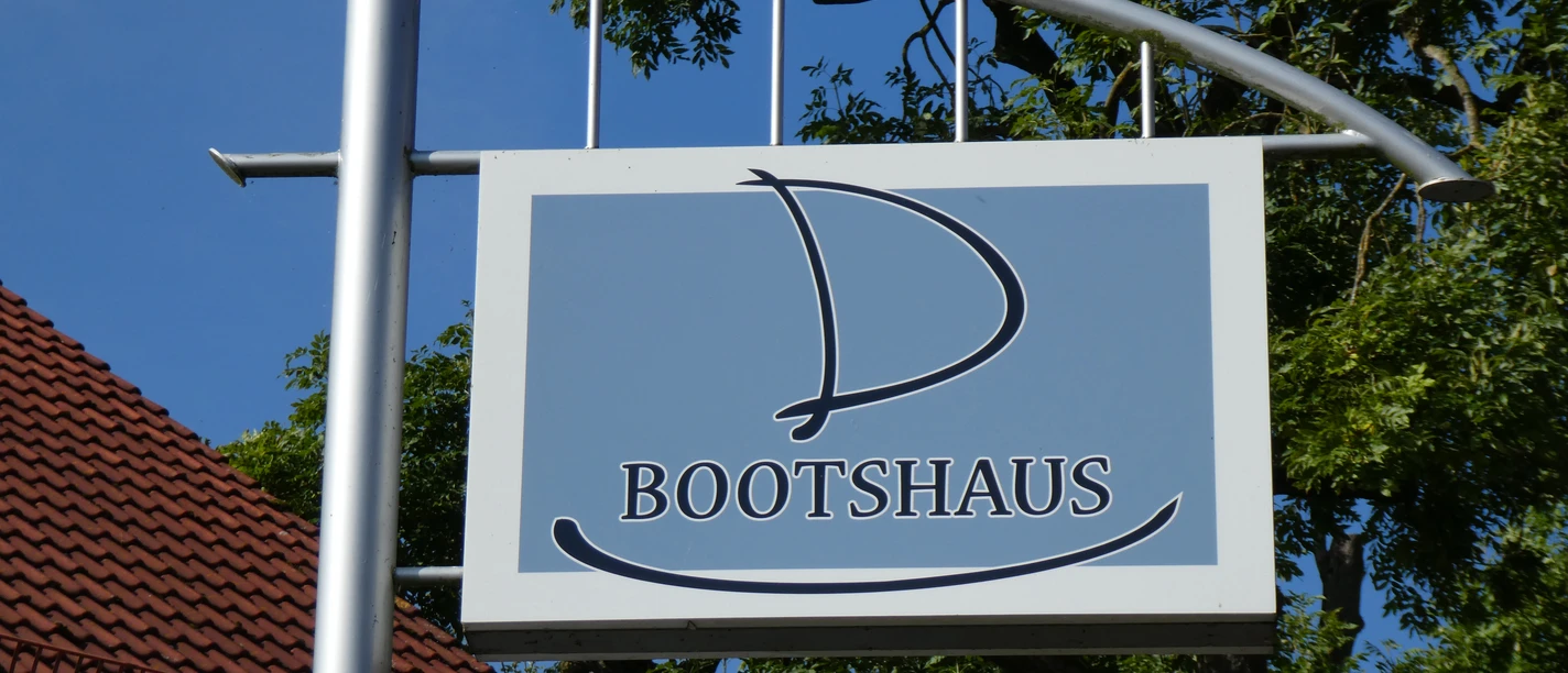 Wooden sign with sailboat-shaped logo of the Bootshaus Hotel-Restaurant in front of green trees.