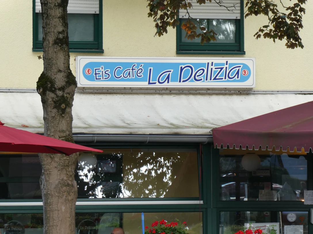 Eiscafé La Delizia Eiscafé La Delizia mit blauem Schild, umgeben von Bäumen und roten Markisen, lädt Besucher ein.Ice cream parlor La Delizia with a blue sign, surrounded by trees and red awnings, invites visitors.Isbaren La Delizia med et blåt skilt, omgivet af træer og røde markiser, indbyder til besøg.IJssalon La Delizia met een blauw uithangbord, omringd door bomen en rode luifels, nodigt bezoekers uit.