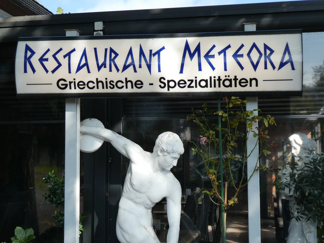 Restaurant Meteora Schild des Restaurants Meteora mit der Aufschrift "Griechische Spezialitäten" und dekorativer Statue.