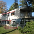 Haus Schminke in Löbau Modernistisches Gebäude mit großen Fenstern, Terrasse und roten Akzenten, von Bäumen umgeben.Modernist building with large windows, terrace and red accents, surrounded by trees.Modernistická budova s velkými okny, terasou a červenými akcenty, obklopená stromy.Modernistyczny budynek z dużymi oknami, tarasem i czerwonymi akcentami, otoczony drzewami.Modernistisch gebouw met grote ramen, terras en rode accenten, omgeven door bomen.Edificio modernista con grandi finestre, terrazza e accenti rossi, circondato da alberi.