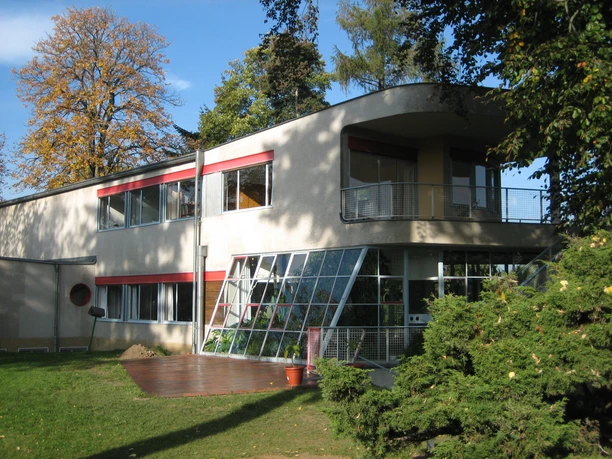 Haus Schminke in Löbau Modernistisches Gebäude mit großen Fenstern, Terrasse und roten Akzenten, von Bäumen umgeben.