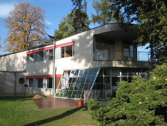 Haus Schminke in Löbau Modernistisches Gebäude mit großen Fenstern, Terrasse und roten Akzenten, von Bäumen umgeben.Modernist building with large windows, terrace and red accents, surrounded by trees.Modernistická budova s velkými okny, terasou a červenými akcenty, obklopená stromy.Modernistyczny budynek z dużymi oknami, tarasem i czerwonymi akcentami, otoczony drzewami.Modernistisch gebouw met grote ramen, terras en rode accenten, omgeven door bomen.Edificio modernista con grandi finestre, terrazza e accenti rossi, circondato da alberi.