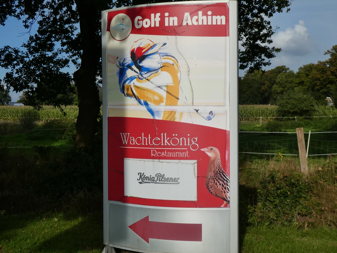 Ein Schild mit der Aufschrift "Golf in Achim" und "Wachtelkönig Restaurant", umgeben von Natur.