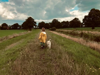 Spaziergang auf dem Deich der Großen Norderbäke Kind im gelben Regenmantel läuft mit weißem Hund auf grünem Deich unter bewölktem Himmel.