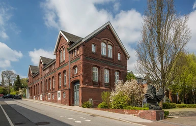 Kattwinkelsche Fabrik Backsteingebäude im historischen Stil mit Satteldach, flankiert von blühenden Bäumen an einer ruhigen Straße.