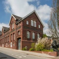 Kattwinkelsche Fabrik Backsteingebäude im historischen Stil mit Satteldach, flankiert von blühenden Bäumen an einer ruhigen Straße.