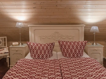 Doppelbett in rustikalem Fachwerkzimmer mit Holzdecke, weißem Kopfteil, rot-gemusterten Kissen und Decken.