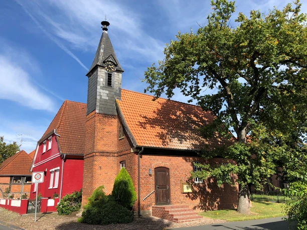 Eine Backsteinkapelle mit Schindelturm und rotem Anbau, umgeben von Bäumen, unter blauem Himmel.
