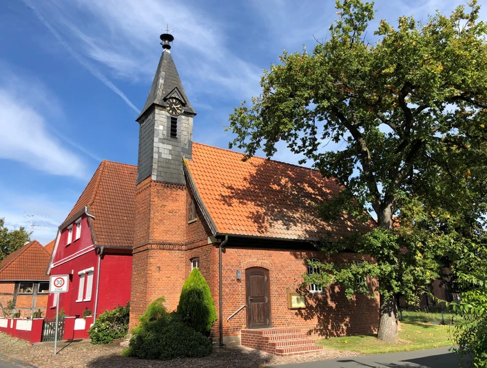 Eine Backsteinkapelle mit Schindelturm und rotem Anbau, umgeben von Bäumen, unter blauem Himmel.