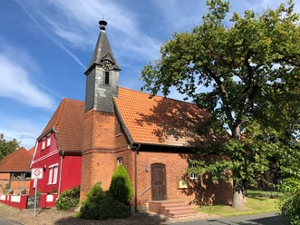 Kapelle Wellie Eine Backsteinkapelle mit Schindelturm und rotem Anbau, umgeben von Bäumen, unter blauem Himmel.