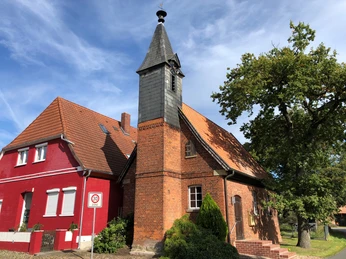 Historische Backsteinkapelle mit Spitzdach und Glockenturm, neben rotem Gebäude, unter blauem Himmel.