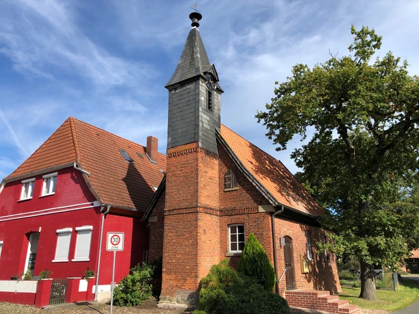 Kapelle Wellie Historische Backsteinkapelle mit Spitzdach und Glockenturm, neben rotem Gebäude, unter blauem Himmel.