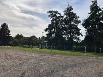 Wohnmobilstellplatz mit Bäumen, Schotterboden und Pollern in einer grünen Umgebung an einem sonnigen Tag.