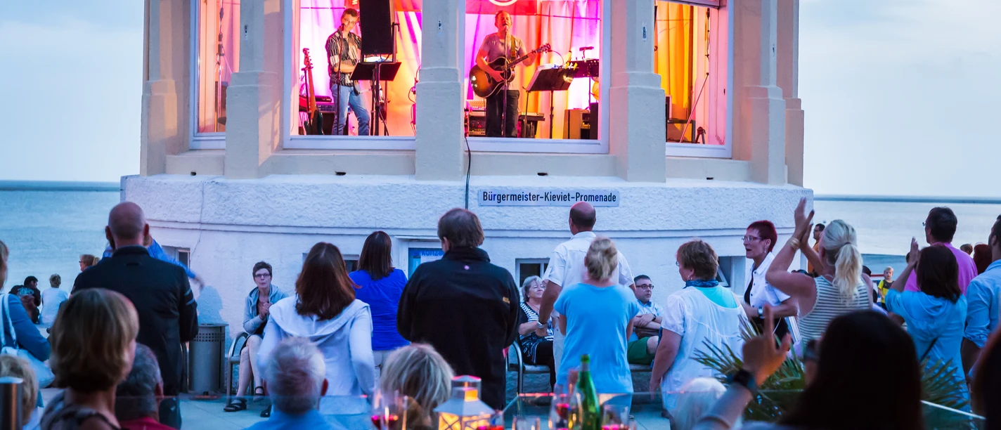 Eine sommerliche Musikveranstaltung mit Liveband in einem Pavillon am Meer zieht begeisterte Zuschauer an.A summer music event with a live band in a pavilion by the sea attracts an enthusiastic audience.