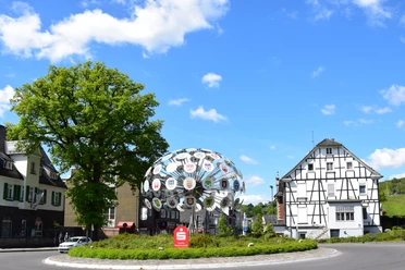 Kunst am Kreisverkehr Kreisverkehr in Bergneustadt mit Fachwerkhaus und moderner Skulptur, blauem Himmel und Bäumen.
