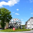 Kunst am Kreisverkehr Kreisverkehr in Bergneustadt mit Fachwerkhaus und moderner Skulptur, blauem Himmel und Bäumen.