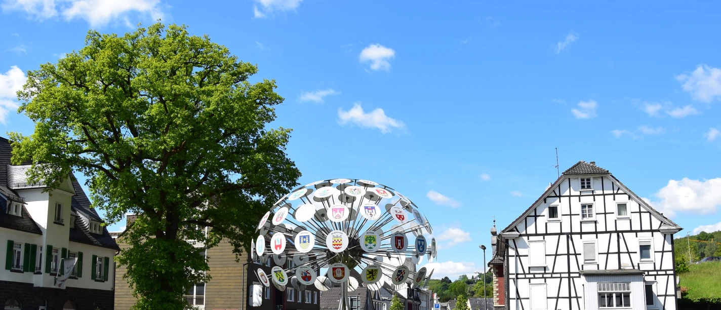 Kunst am Kreisverkehr Kreisverkehr in Bergneustadt mit Fachwerkhaus und moderner Skulptur, blauem Himmel und Bäumen.
