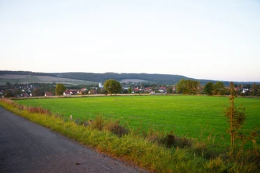 Grüne Wiesen führen zu einem ruhigen Dorf mit verstreuten Häusern, umgeben von sanften Hügeln.
