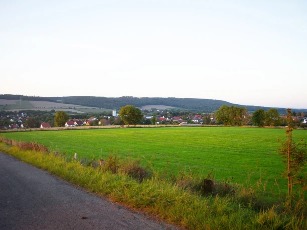 Grüne Wiesen führen zu einem ruhigen Dorf mit verstreuten Häusern, umgeben von sanften Hügeln.