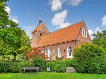 st-johannes-kirche-bad-zwischenahn.jpg Backsteinkirche St. Johannes mit rotem Satteldach und Turm, umgeben von grünen Bäumen unter blauem Himmel.