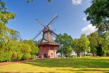 muehle-kurpark-freilichtmuseum-sommer.jpg Historische Windmühle im Kurpark, umgeben von grünen Bäumen unter blauem Sommerhimmel.