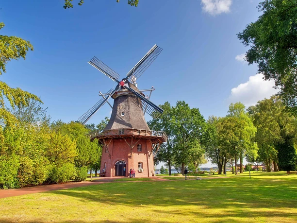 muehle-kurpark-freilichtmuseum-sommer.jpg Historisk vindmølle i spa-haven, omgivet af grønne træer under en blå sommerhimmel.